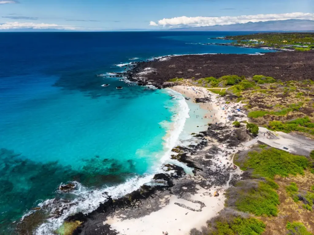 Manini’owali Beach on the Big Island of Hawaii, a small white-sand beach with clear turquoise waters, black lava rocks, and gentle waves, ideal for swimming and snorkeling.