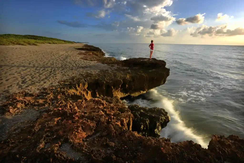 A person stands on a rocky outcrop overlooking the ocean at sunset, with waves crashing below and a sandy beach extending into the distance under a partly cloudy sky.