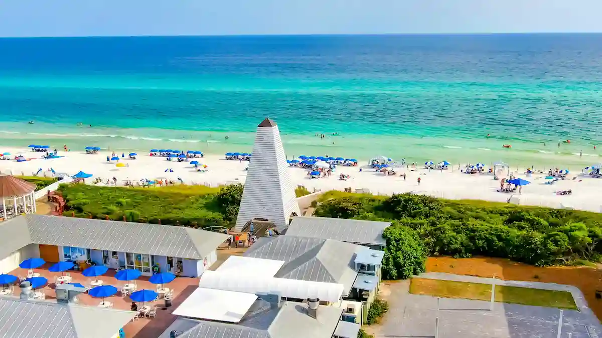 Aerial view of a beach with turquoise water, white sand, people relaxing under umbrellas, and a white lighthouse surrounded by buildings and greenery in the foreground.