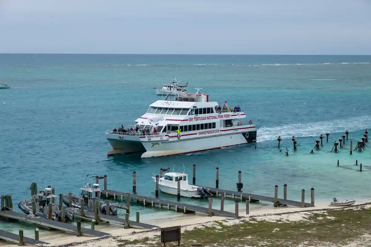 A white passenger ferry approaches a dock on turquoise water, with people visible on board. A small boat is tied to the dock, and empty wooden posts extend into the sea under a cloudy sky.