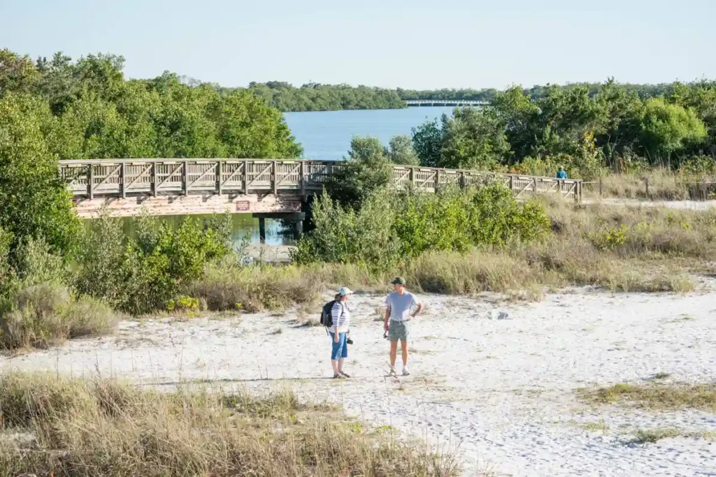 Two people stand on a sandy path near green brush, with a wooden bridge and a river or lake in the background under a clear sky.