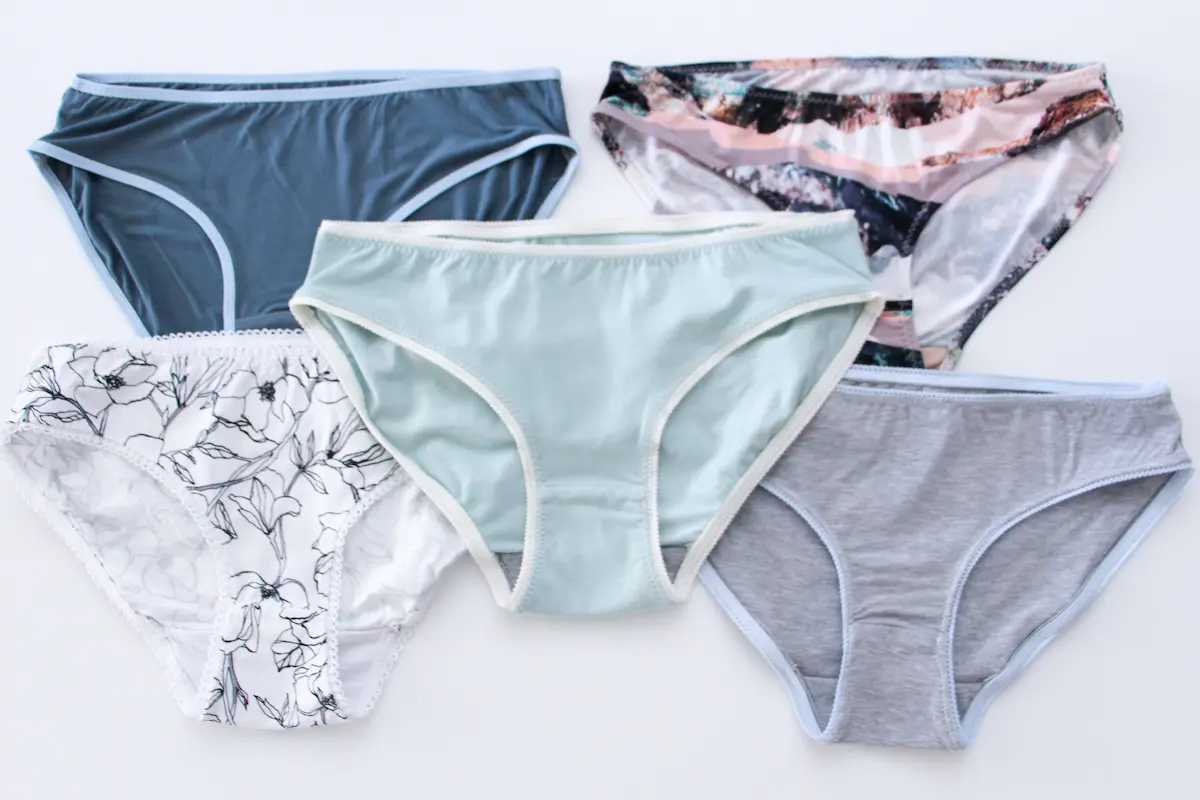 Five pairs of womens panties in different colors and patterns, including blue, mint green, grey, white with a floral design, and a multicolored pattern, arranged in a flat lay on a white surface.