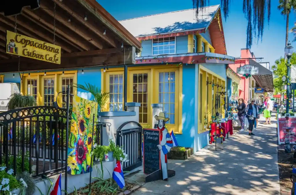 A vibrant street scene shows a bright blue and yellow Cuban café with people walking by, colorful decorations, and flags, under a clear blue sky.