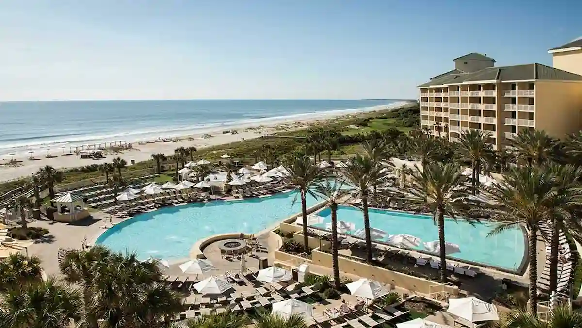 A beachfront resort with a large outdoor pool surrounded by palm trees and lounge chairs, adjacent to a sandy beach and ocean, with a multi-story hotel building on the right under a clear blue sky.