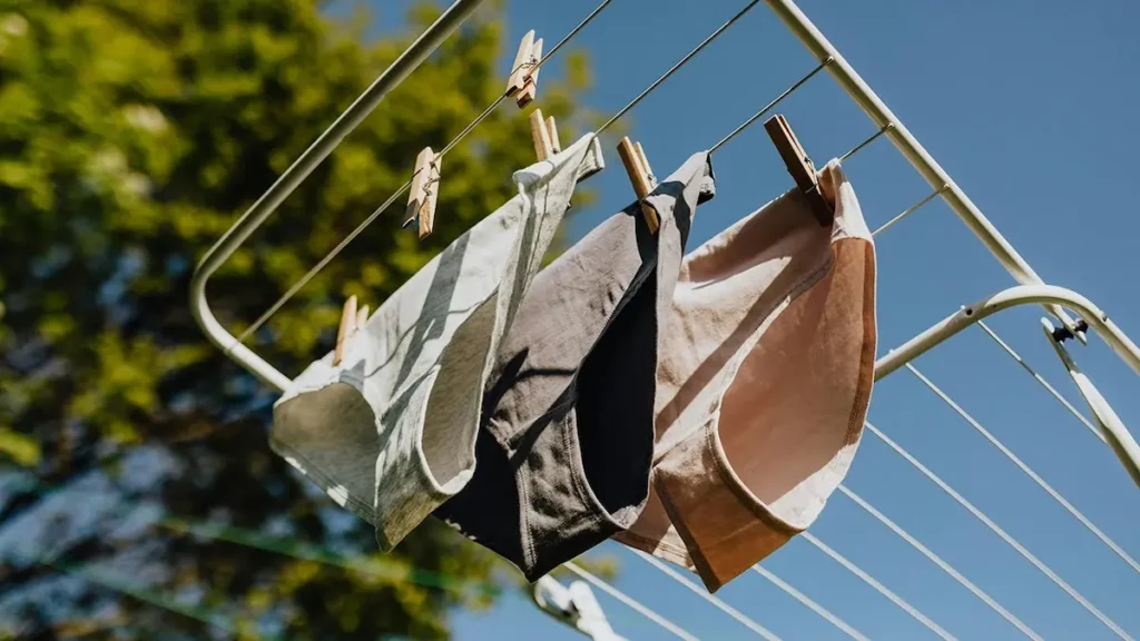 Three pairs of underwear in different colors hanging on a clothesline with clothespins, drying in the sunlight against a blue sky and blurred greenery in the background.