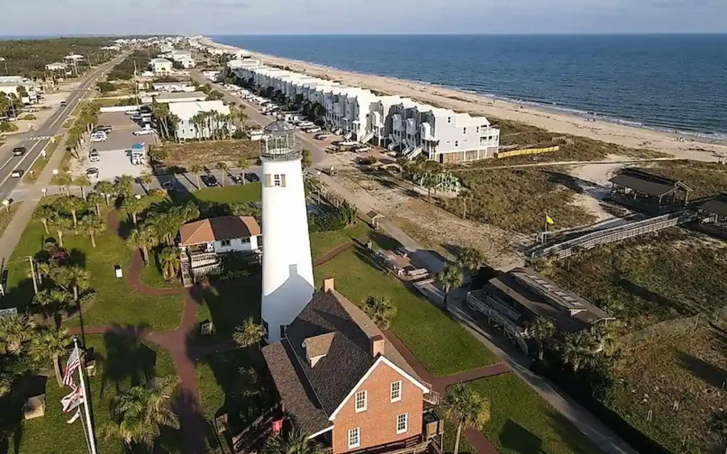 Aerial view of a white lighthouse near the coast, surrounded by a few buildings, with a sandy beach and ocean to the right and a road with houses to the left, under a clear blue sky.