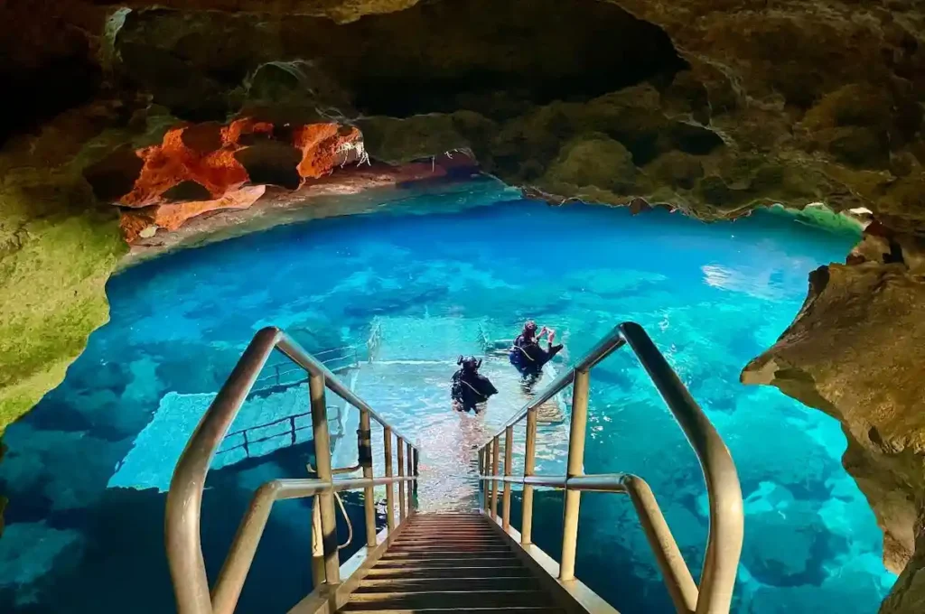 A metal staircase leads down into a vibrant blue underground cave pool, where several scuba divers are preparing to enter the water surrounded by rocky cave walls.