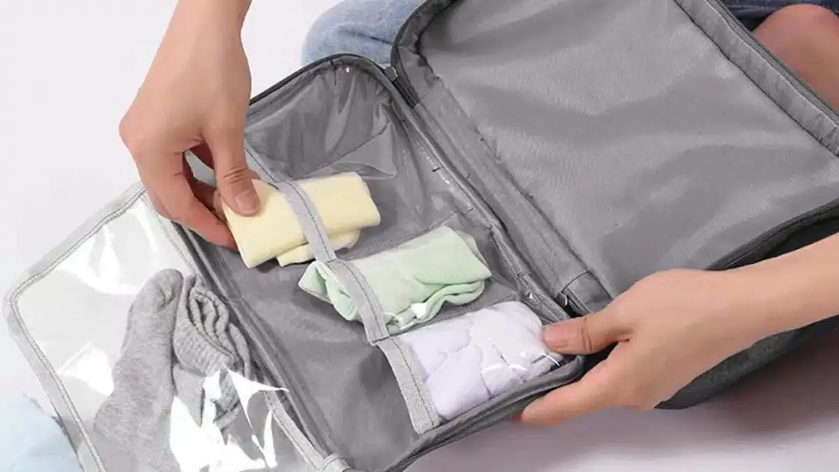 A person organizes small clothing items, such as socks and underwear, into clear zippered compartments of a gray travel organizer bag.