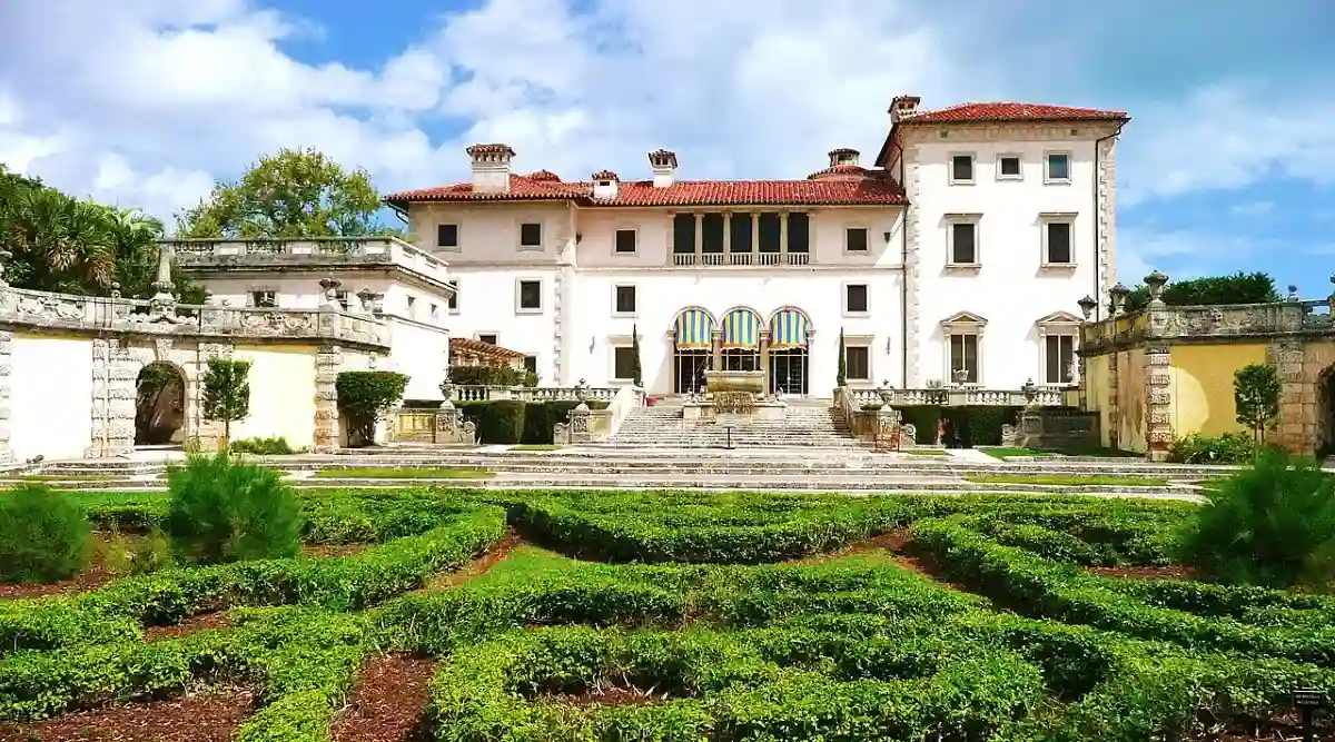 A large, elegant villa with white walls, red-tiled roof, and arched windows stands behind a neatly manicured formal garden with geometric hedges, under a partly cloudy sky.