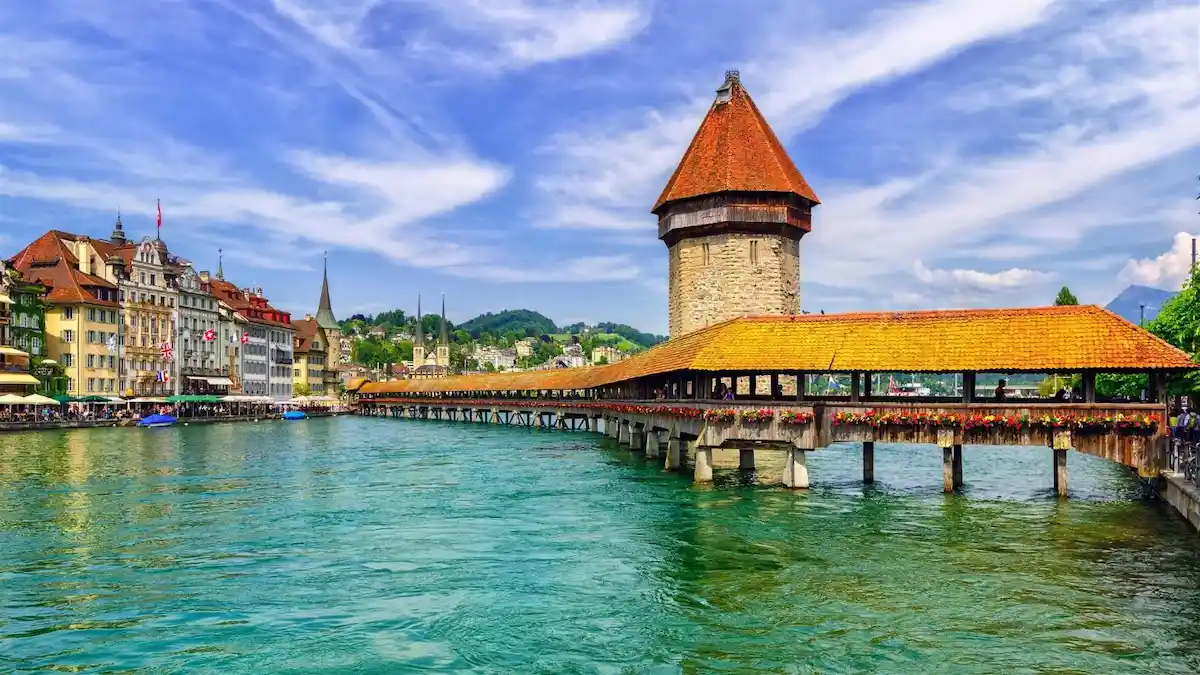 A covered wooden bridge with a stone tower crosses a turquoise river, with colorful buildings and green hills in the background under a vibrant blue sky.