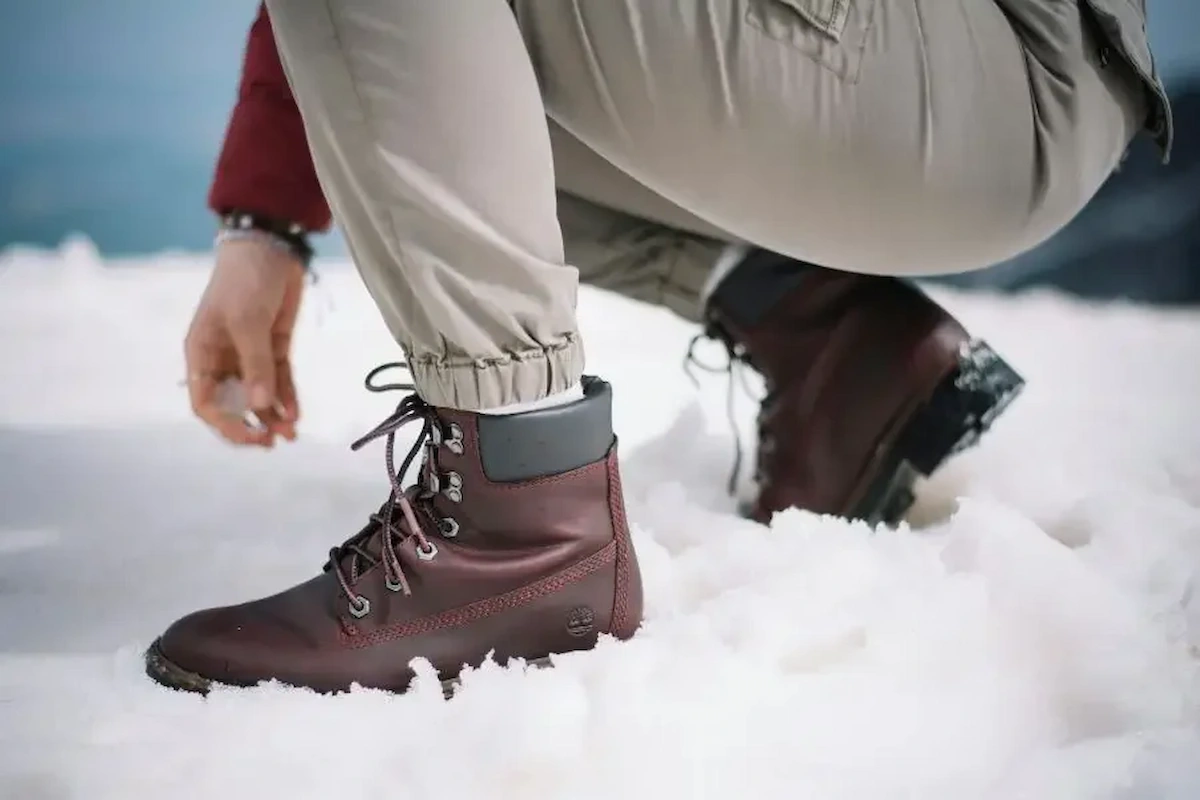 A person wearing brown hiking boots and light-colored pants crouches in the snow, adjusting a pant leg.