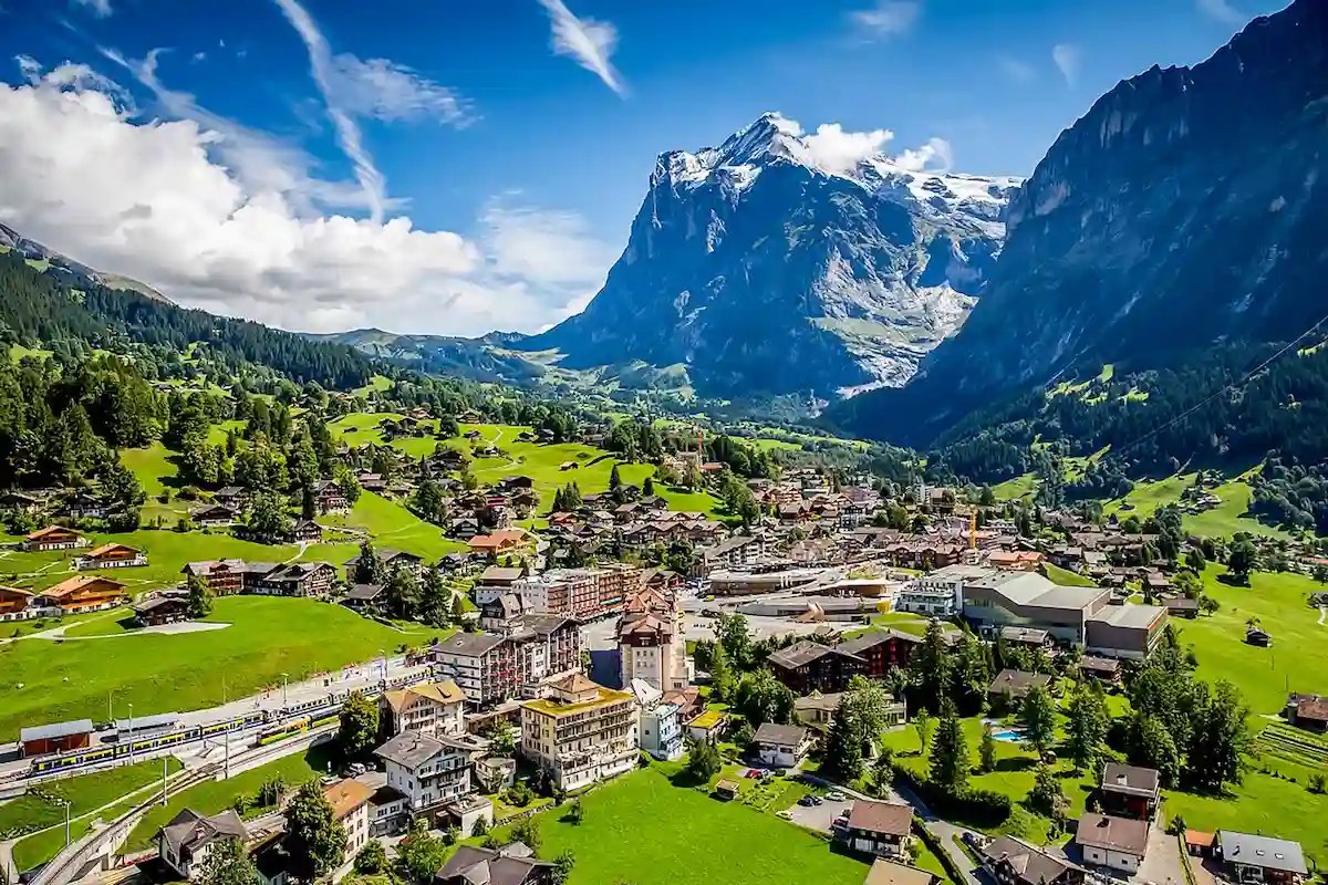 A picturesque alpine village with scattered houses and buildings, surrounded by lush green fields and dense trees, sits at the base of towering, snow-capped mountains under a bright blue sky with scattered clouds.