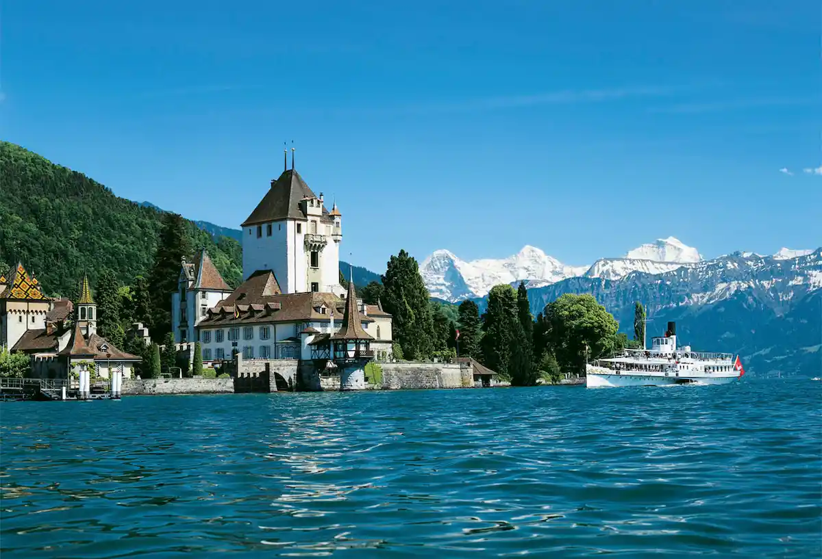 A scenic view of a lakeside castle with a white boat nearby, set against green hills and snow-capped mountains under a clear blue sky.