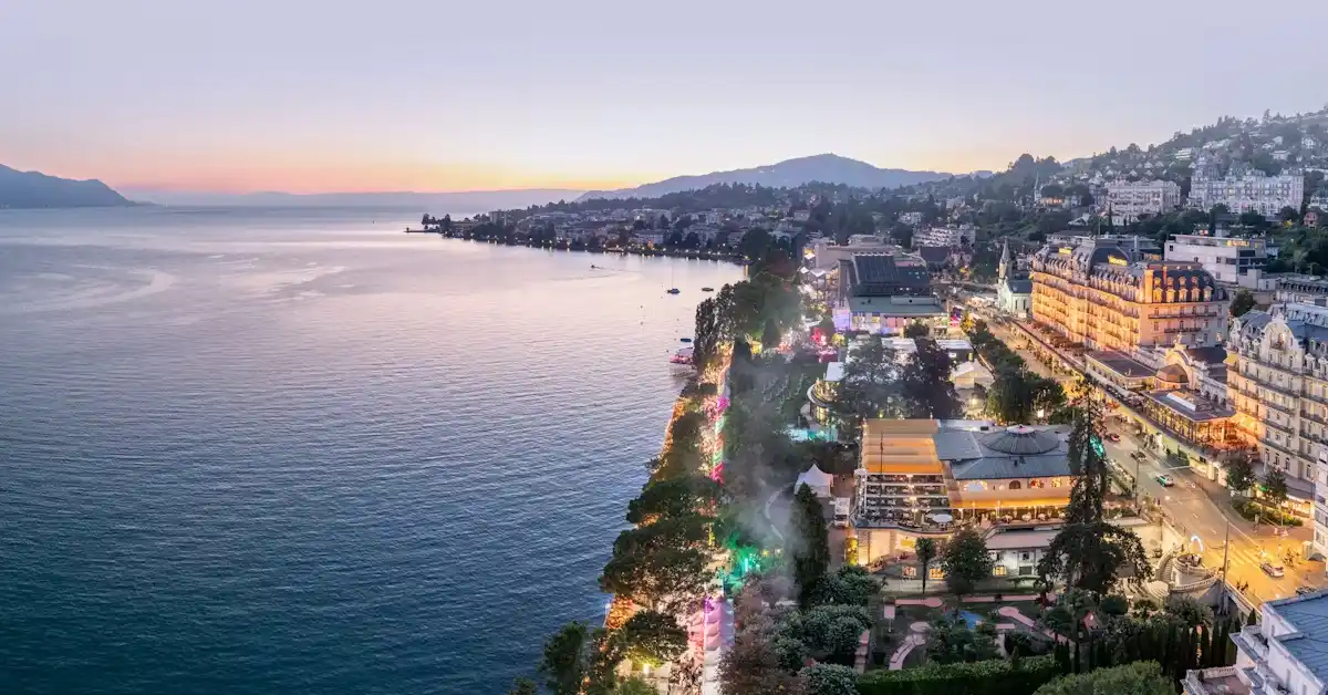 Aerial view of a coastal city at sunset, with calm water on the left and brightly lit buildings, streets, and hills on the right. The sky is pastel-colored, and boats are visible near the shore.