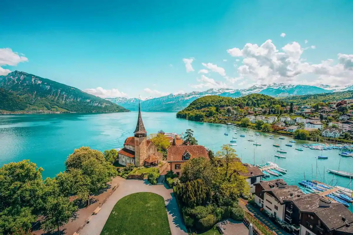 A scenic lakeside village with a pointed church steeple, surrounded by trees, mountains, and boats docked along the waterfront under a bright blue sky.