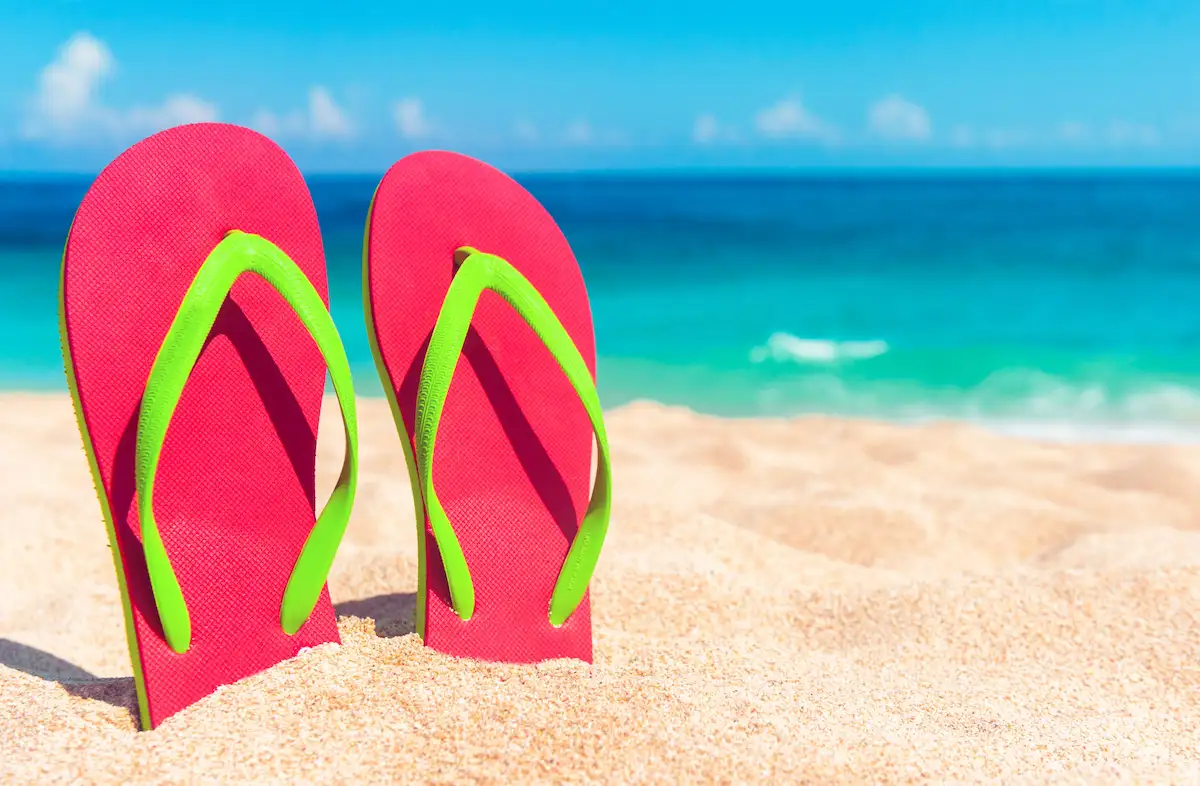 A pair of bright pink flip-flops with green straps standing upright in the sand on a sunny beach, with turquoise ocean waves and a blue sky in the background.