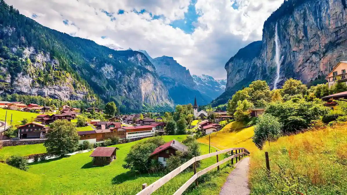 A scenic mountain village with wooden houses, lush green meadows, tall cliffs, and a waterfall cascading down rocks under a partly cloudy sky.