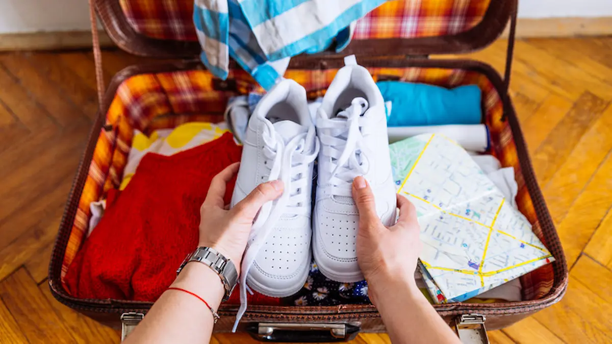 A person places a pair of white sneakers into an open suitcase packed with clothes, a map, and other travel essentials on a wooden floor.