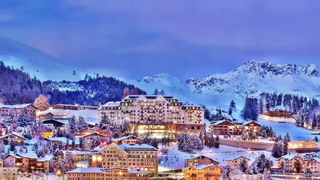 A picturesque winter scene of a snow-covered mountain village, with brightly lit buildings nestled among pine trees and majestic mountains in the background under a purple-tinged evening sky.