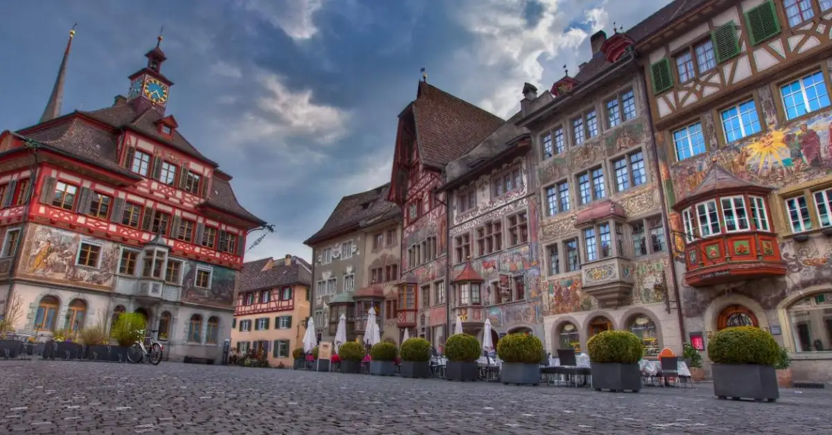 Colorful, ornate historic buildings with detailed frescoes line a cobblestone square under a partly cloudy sky, with potted plants and empty café tables in the foreground.