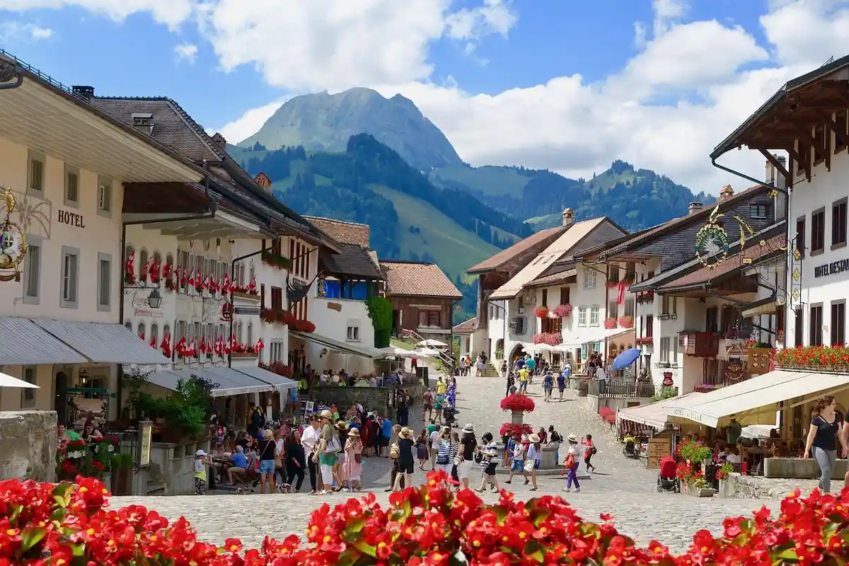 A bustling cobblestone street in a charming alpine village, lined with white buildings and red flowers, with people walking and dining outdoors. Green mountains rise in the background under a partly cloudy sky.