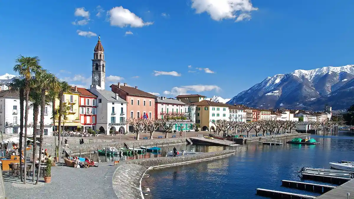 Colorful buildings line a waterfront promenade with outdoor seating, palm trees, and a church tower, set against snow-capped mountains under a bright blue sky.