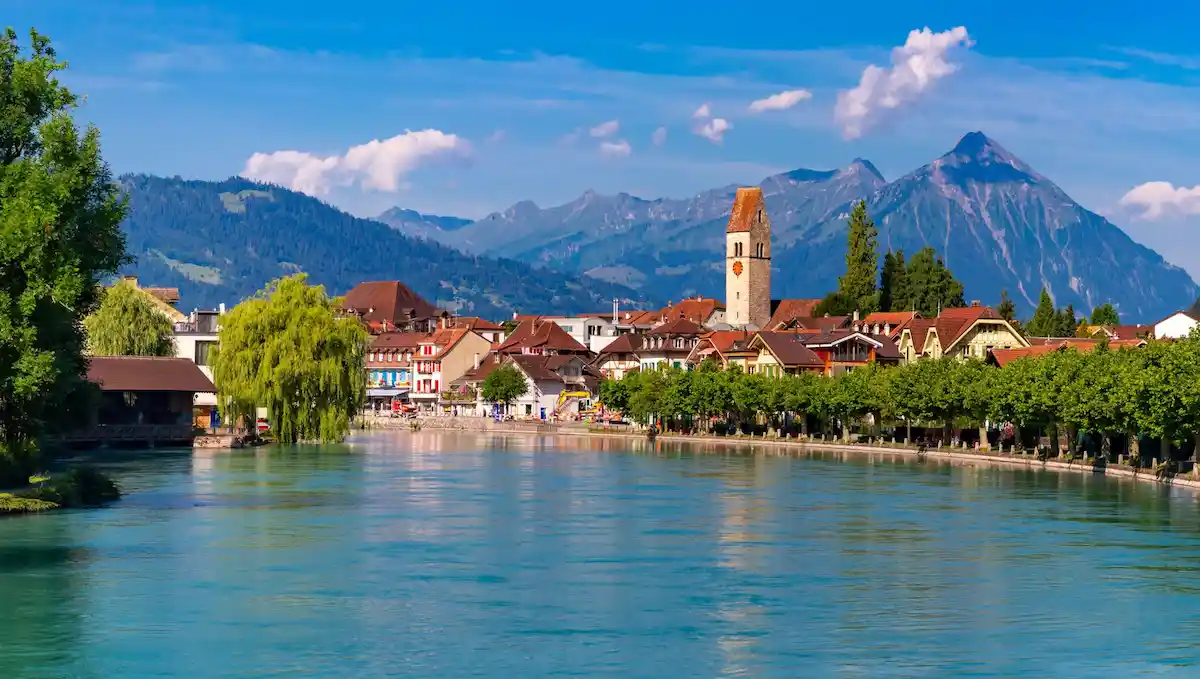 A scenic lakeside town with red-roofed buildings, a church tower, lush green trees, and mountains in the background under a blue sky with some clouds.