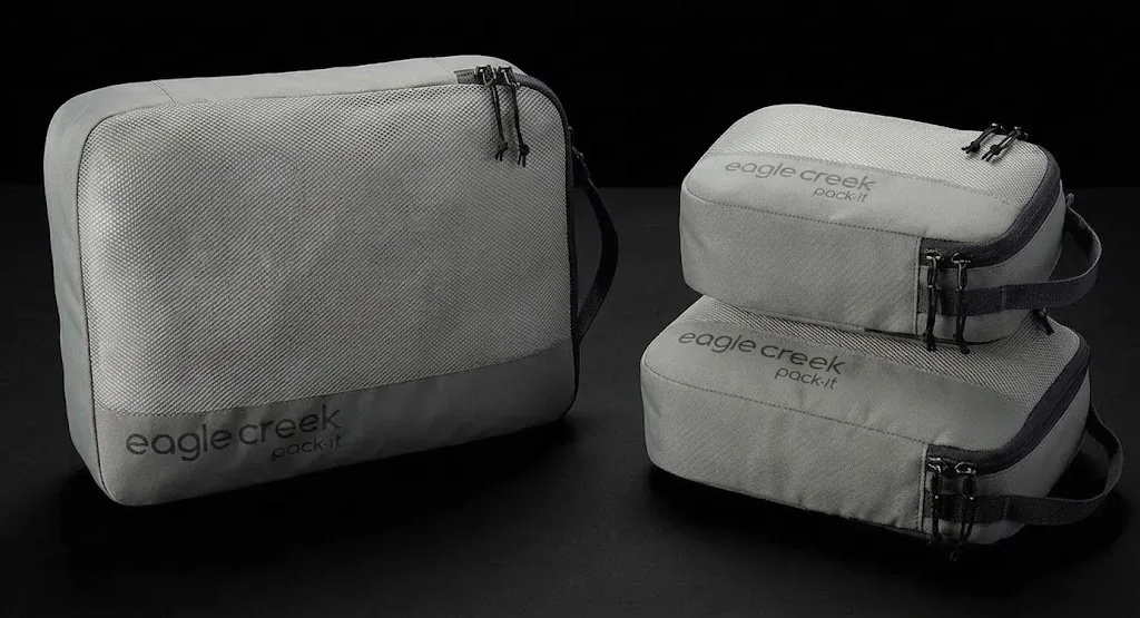 Three grey Eagle Creek Pack-It travel organizer bags are displayed against a black background. One bag is larger and rectangular, while the other two are smaller, stacked, and more compact in shape.