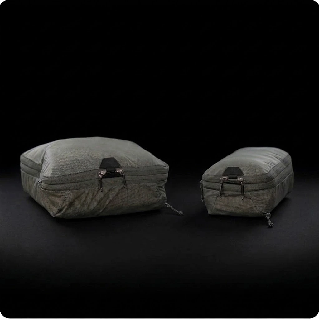 Two gray, zippered packing cubes sit side by side against a black background; one cube is packed full and bulky, while the other is compressed and much slimmer.